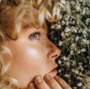 An attractive blonde woman lightly touches her face which is close to a wall of flowers
